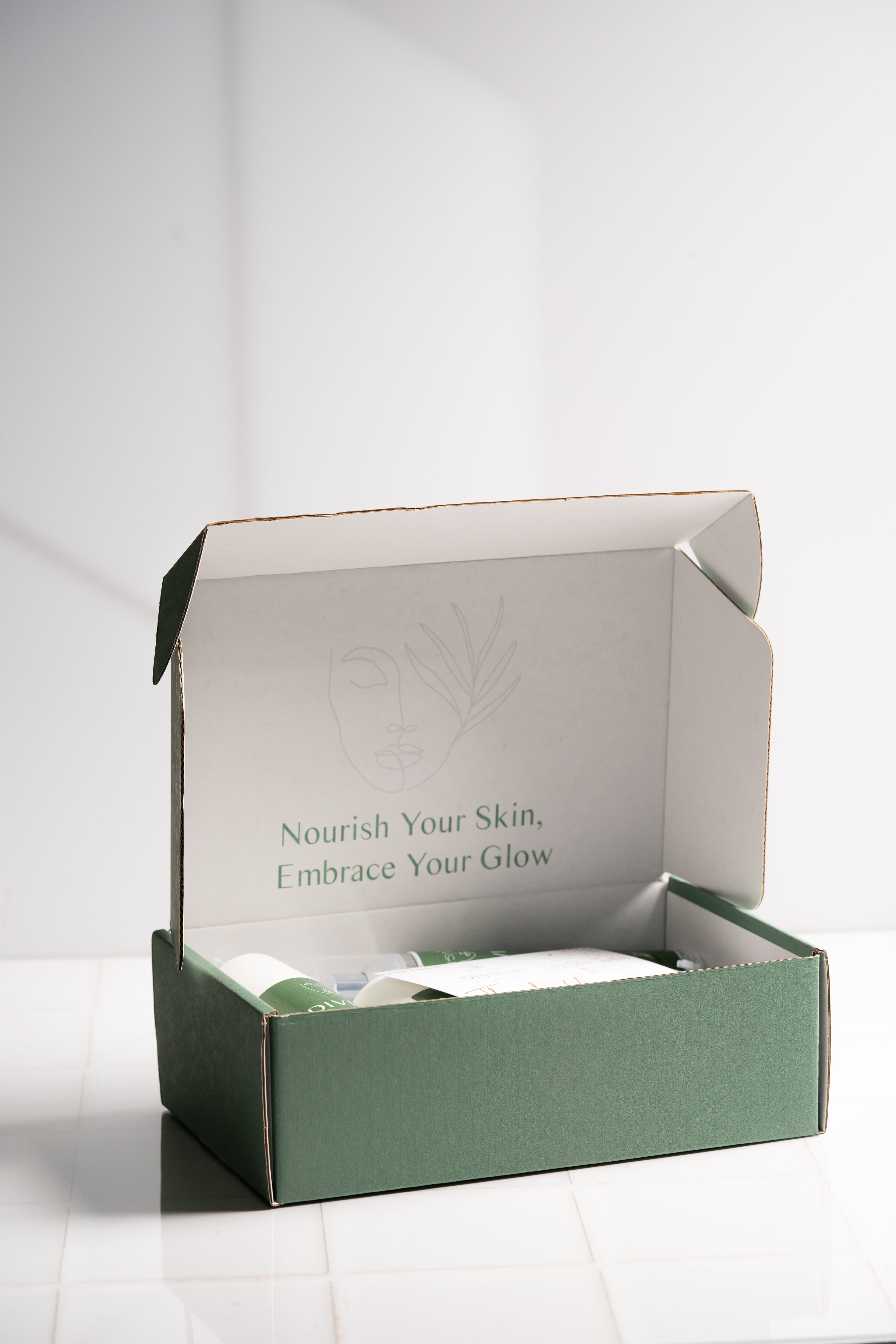 A green and white open box with skincare products inside sits on a white tiled surface under bright lighting. The inside lid reads, "Nourish Your Skin, Embrace Your Glow," with a minimalist line drawing of a face and leaf. This Divina Natural Divibox contains 7 products, now available at a reduced price.