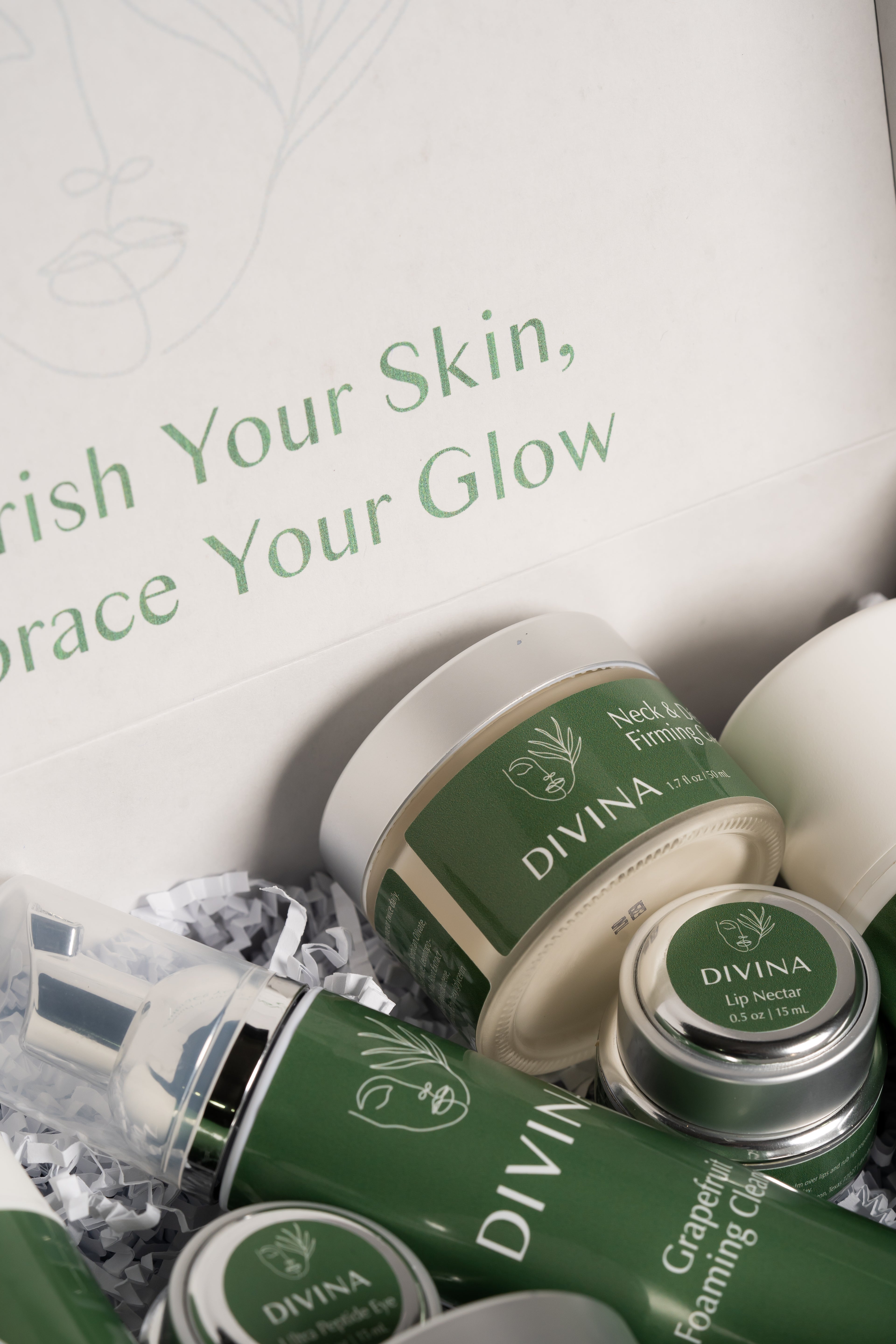 A close-up of the Divibox skincare set from Divina Natural. The set includes a neck firming cream, lip nectar, and grapefruit foaming cleanser, all packaged in a box with the text "Nourish Your Skin, Embrace Your Glow." The products feature green labels with a white leaf logo. Act now for this reduced price!