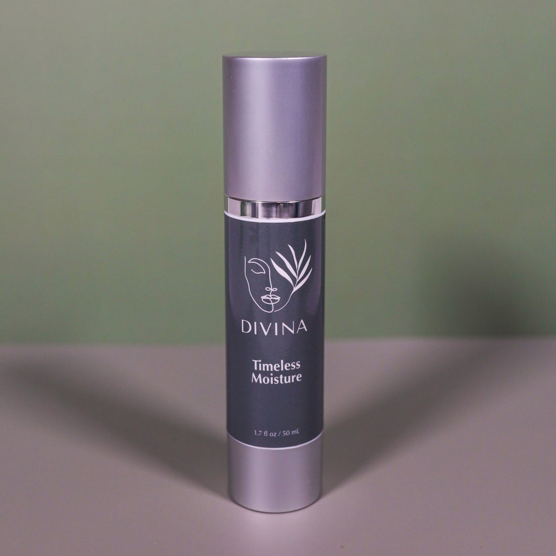 A silver and dark gray bottle of Divina Timeless Moisture stands upright on a gray surface against a green background, displaying white text and a minimalist plant logo.