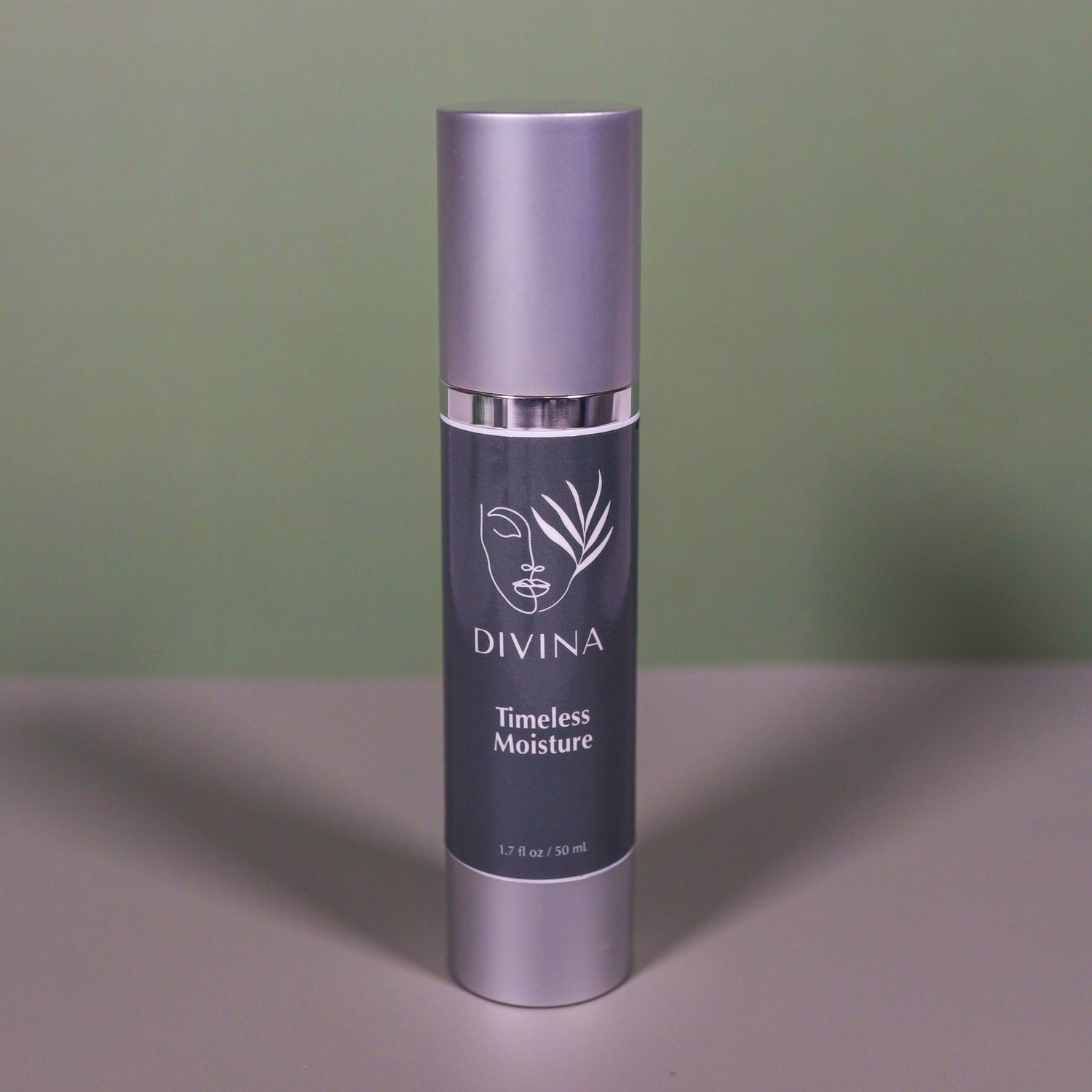 A silver and dark gray bottle of Divina Timeless Moisture stands upright on a gray surface against a green background, displaying white text and a minimalist plant logo.