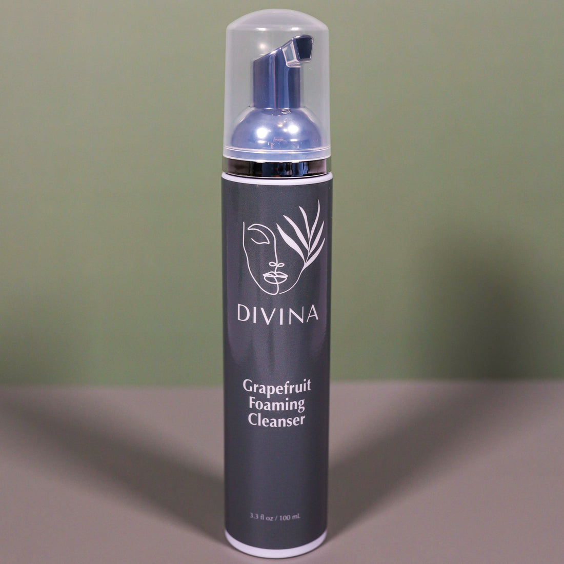 A bottle of Divina Grapefruit Foaming Cleanser with a clear cap, gray label, and white text stands upright against a green and gray background.