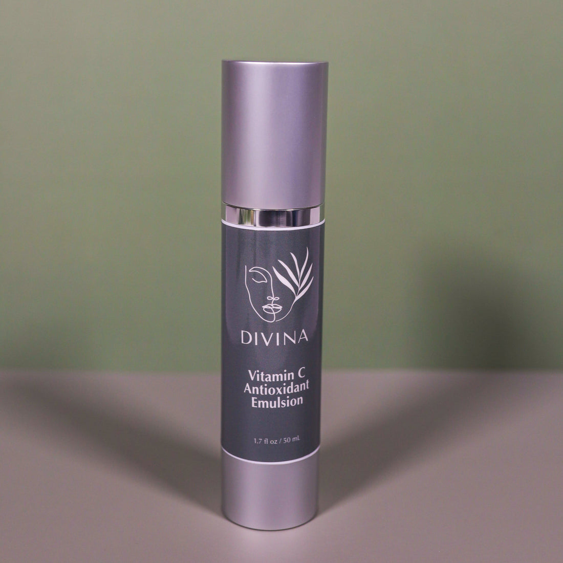 A silver and gray bottle labeled “Divina Vitamin C Antioxidant Serum,” designed for anti-aging, stands upright on a light surface against a muted green background. The label has a minimalist face and leaf design.