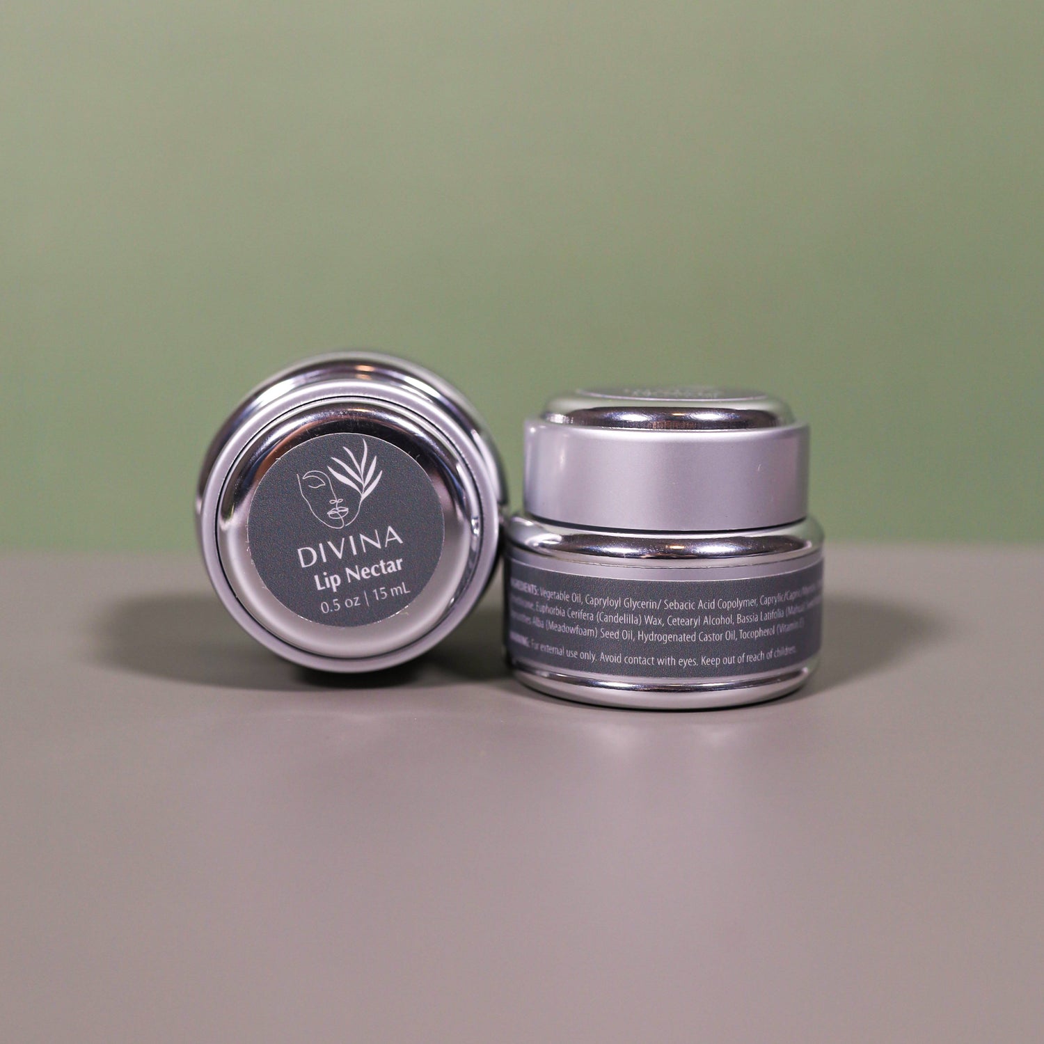 Two small silver jars of Divina’s Lip Nectar, a luxurious lip treatment, sit against a neutral background—one upright displaying the side label, the other tilted to spotlight the lid and Divina branding.